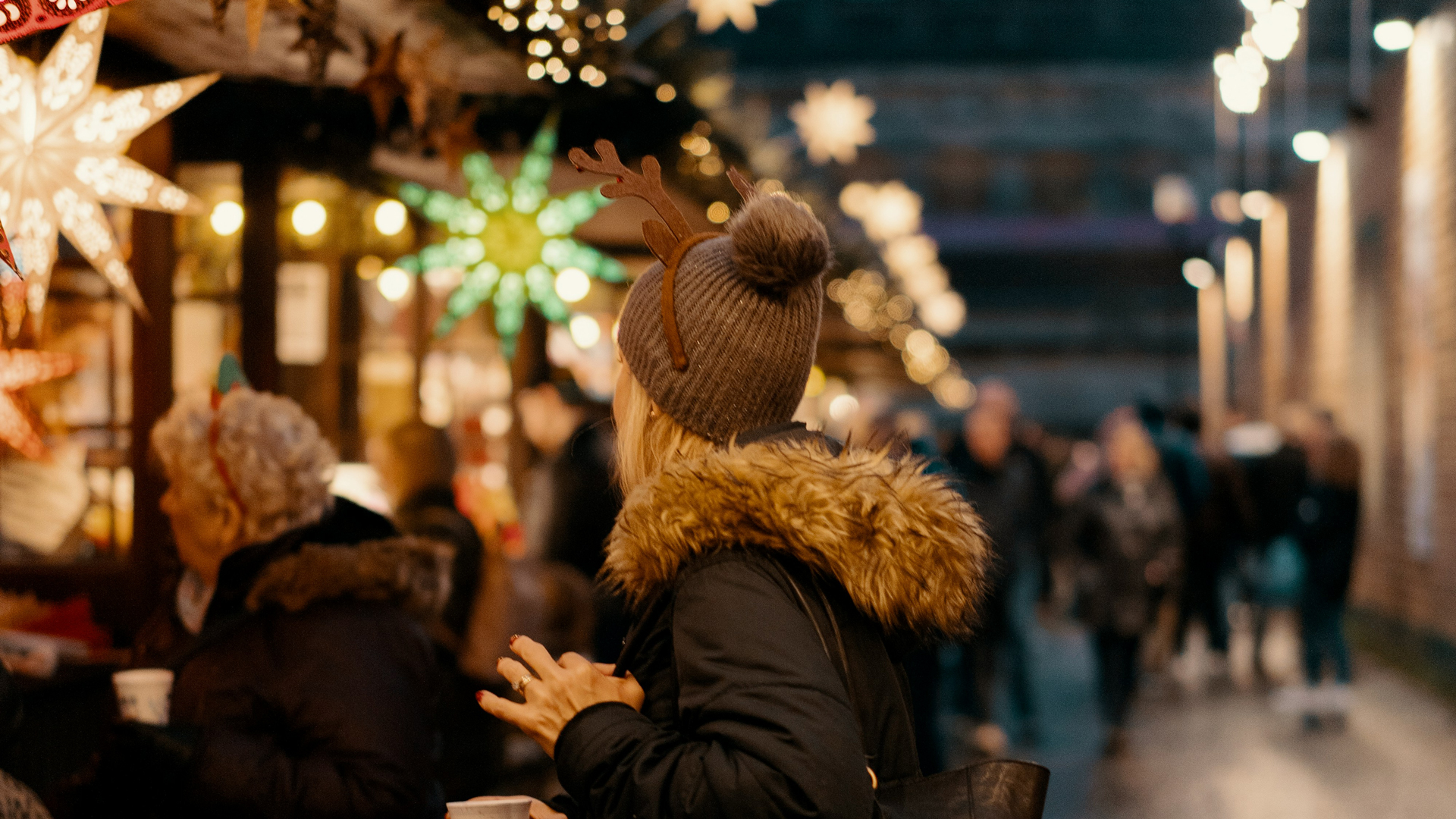 An image of a woman with a winter coat, hat and reindeer ears looking away from the camera. The background is out of focus but shows Christmas lights,