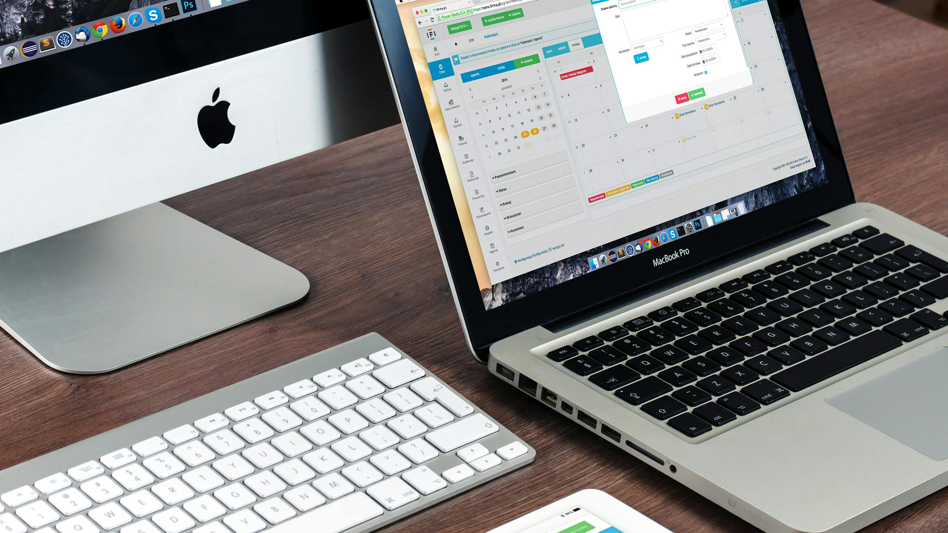 Laptop and desktop computer on a wooden desk with a keyboard in front.