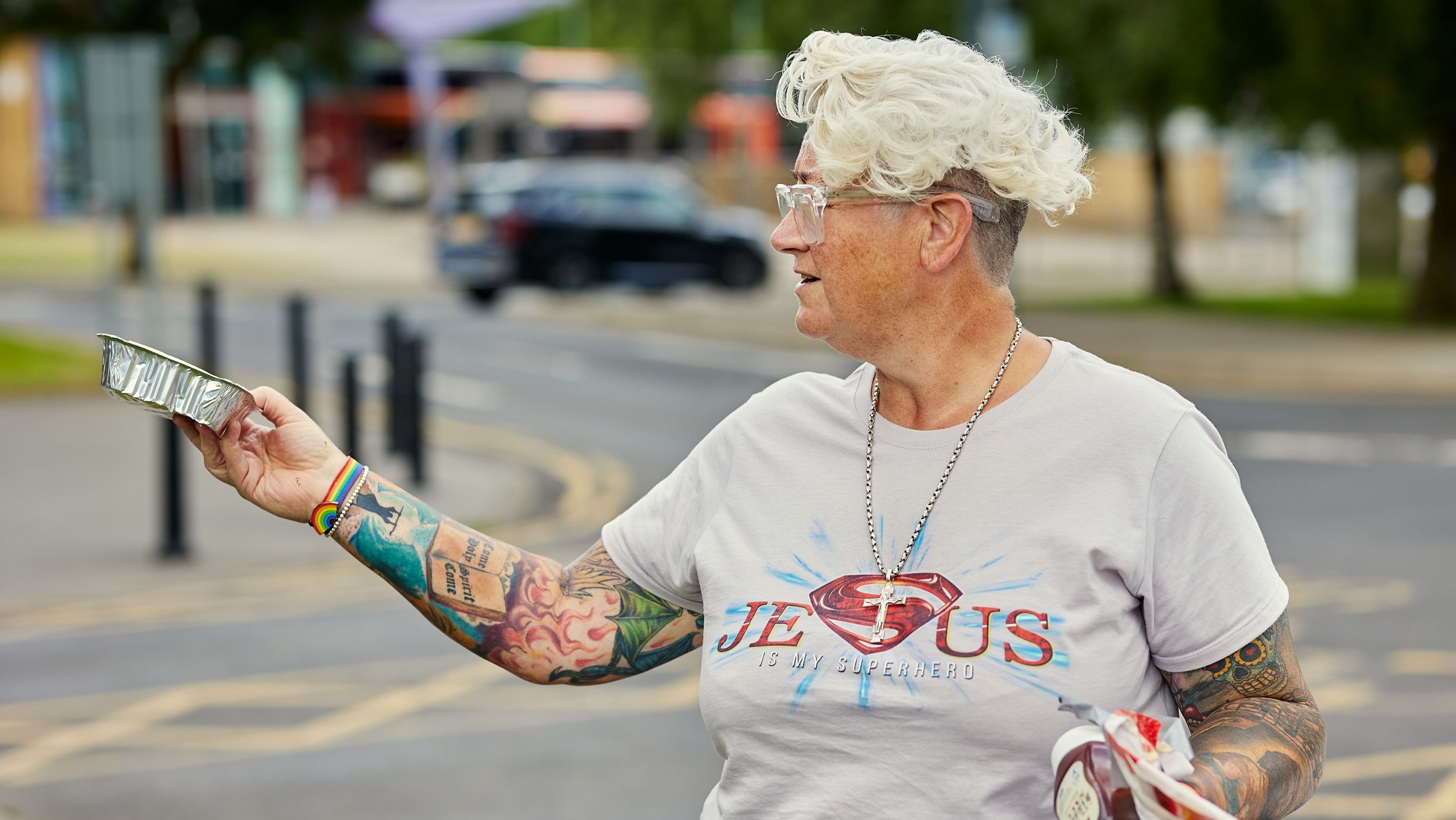 Person with short white hair and tattoos holds a foil food tray while standing on a street.