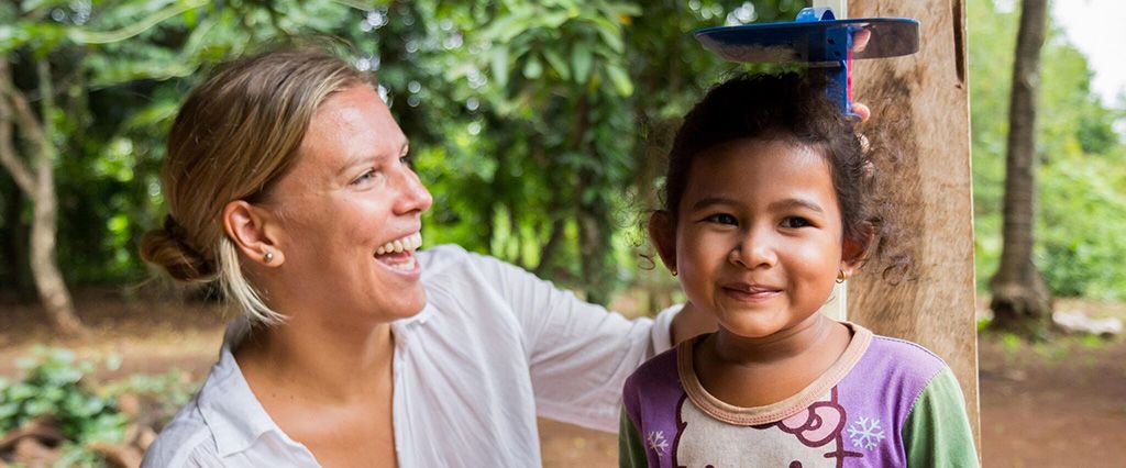 A woman laughs while standing next to a smiling young girl in a wooded outdoor setting.