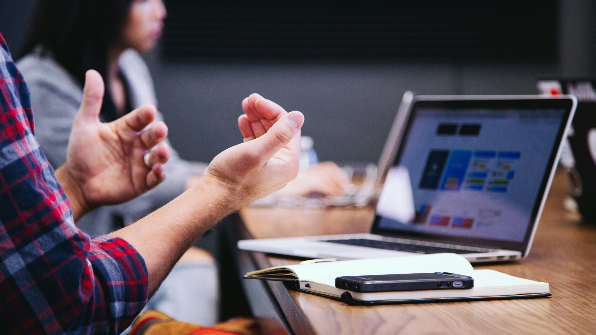 An image of a laptop screen on a desk. A pair of hands positioned as if talking in a meeting.